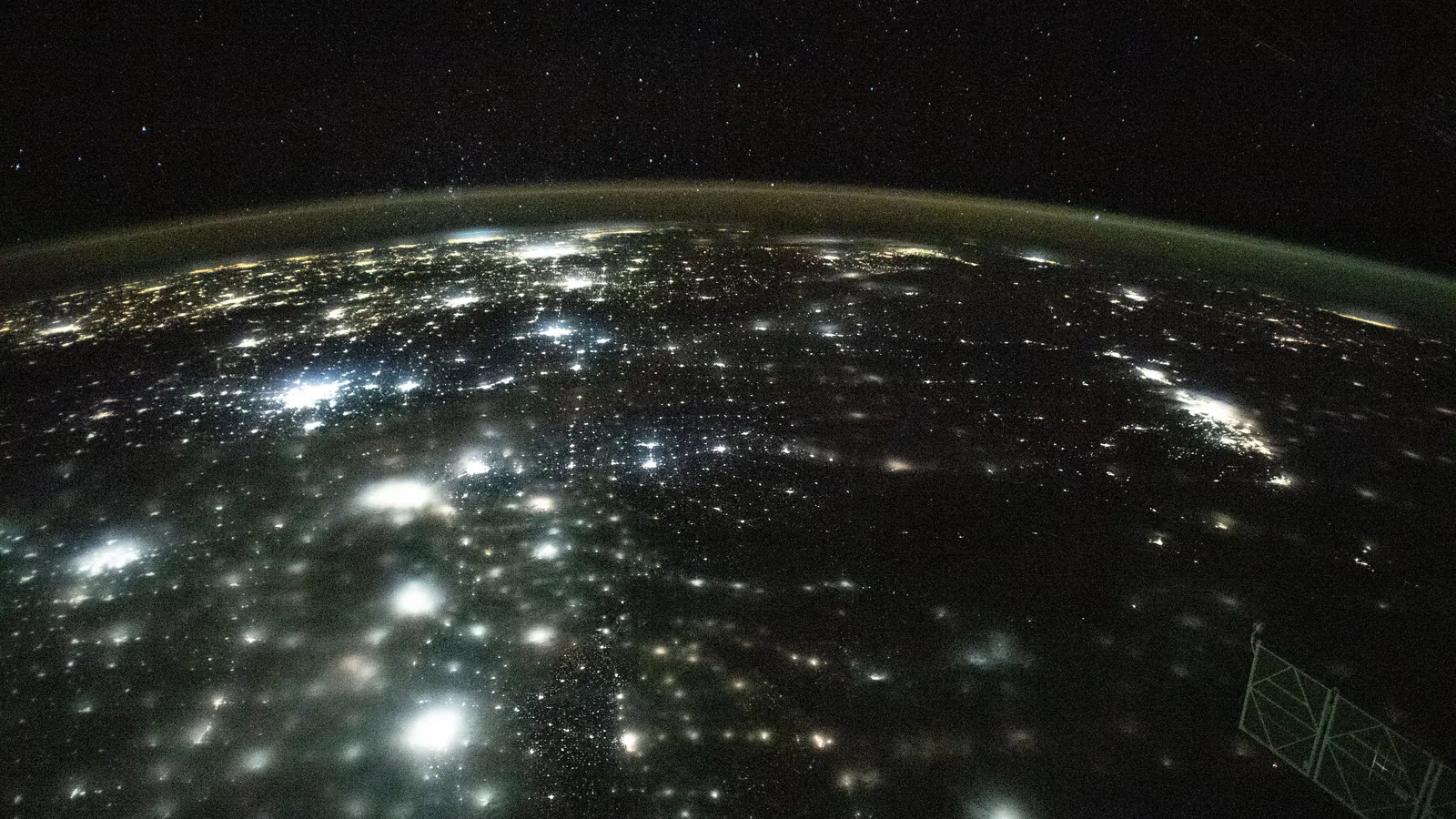 Clusters of city lights spread across a continent at night under a thin green band of airglow, photographed from the International Space Station