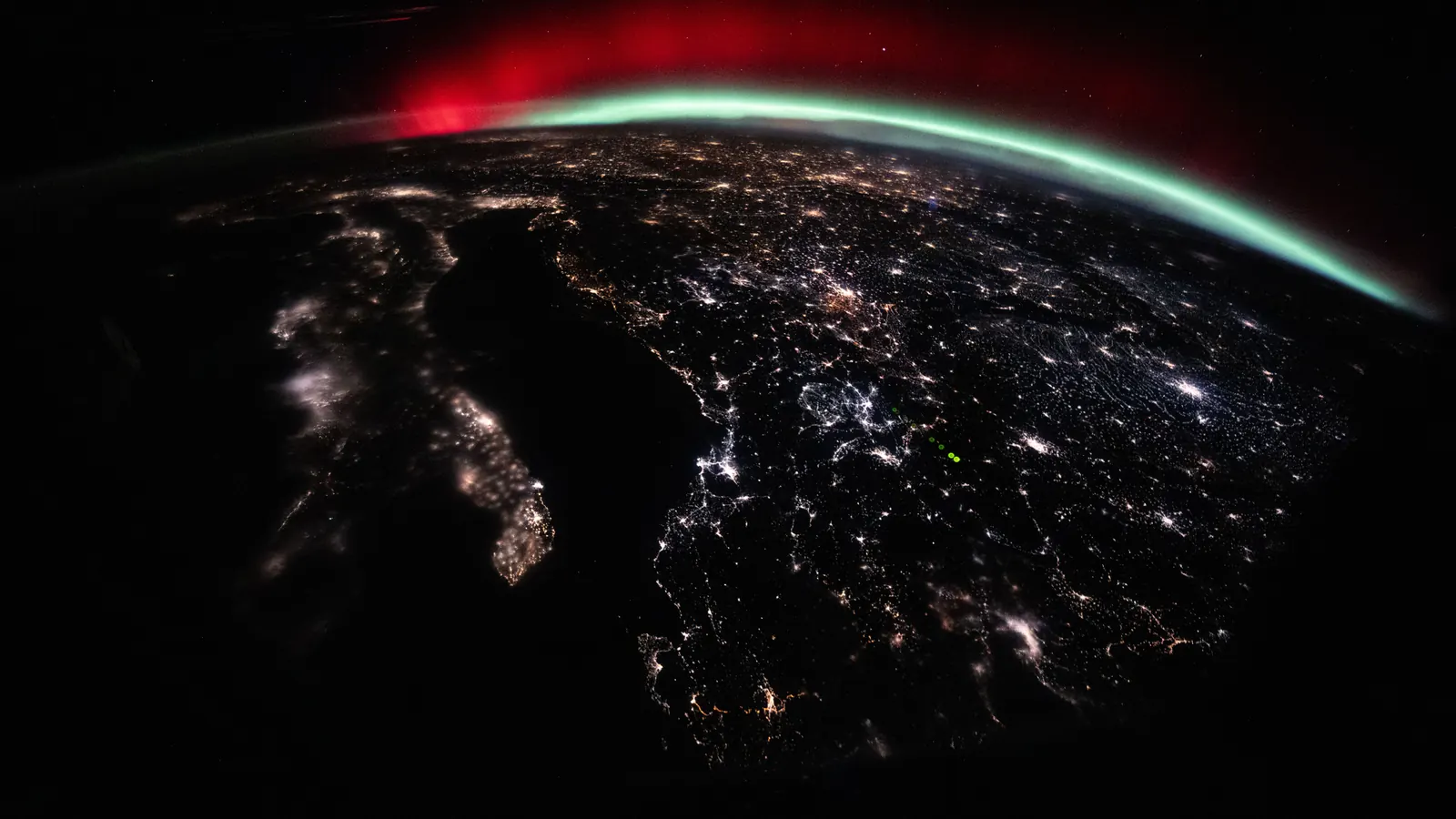 A green and red aurora arcing along the horizon above the city lights of Italy and central Europe, photographed from the International Space Station on January 19, 2026