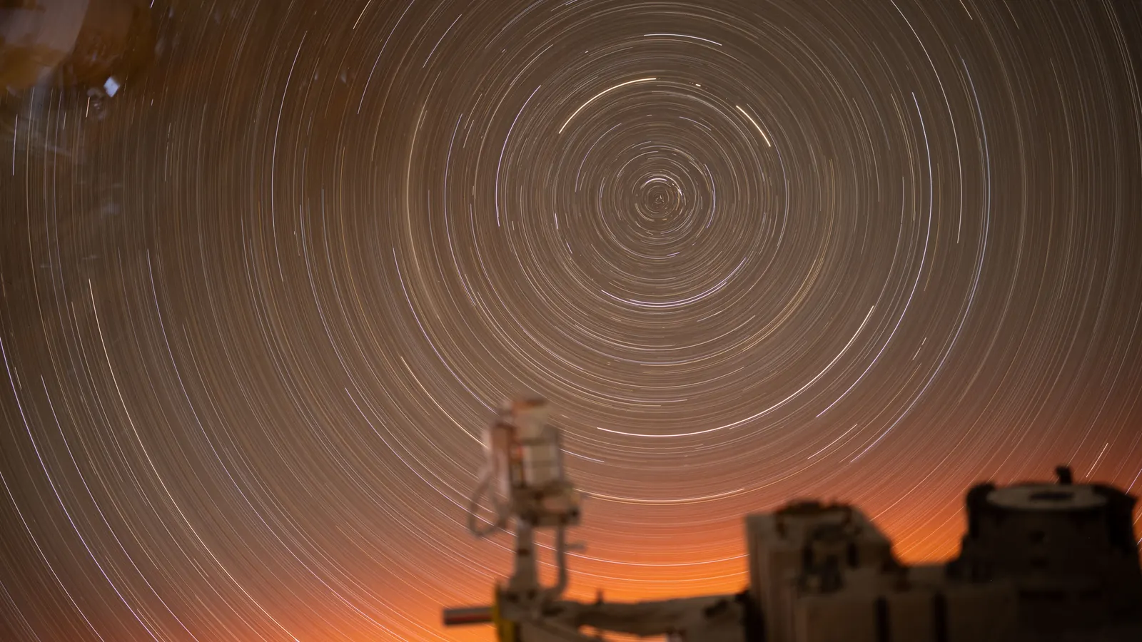 Stars drawn into circular arcs in a long exposure taken from the International Space Station, with the station's hardware lit orange by the sunrise below