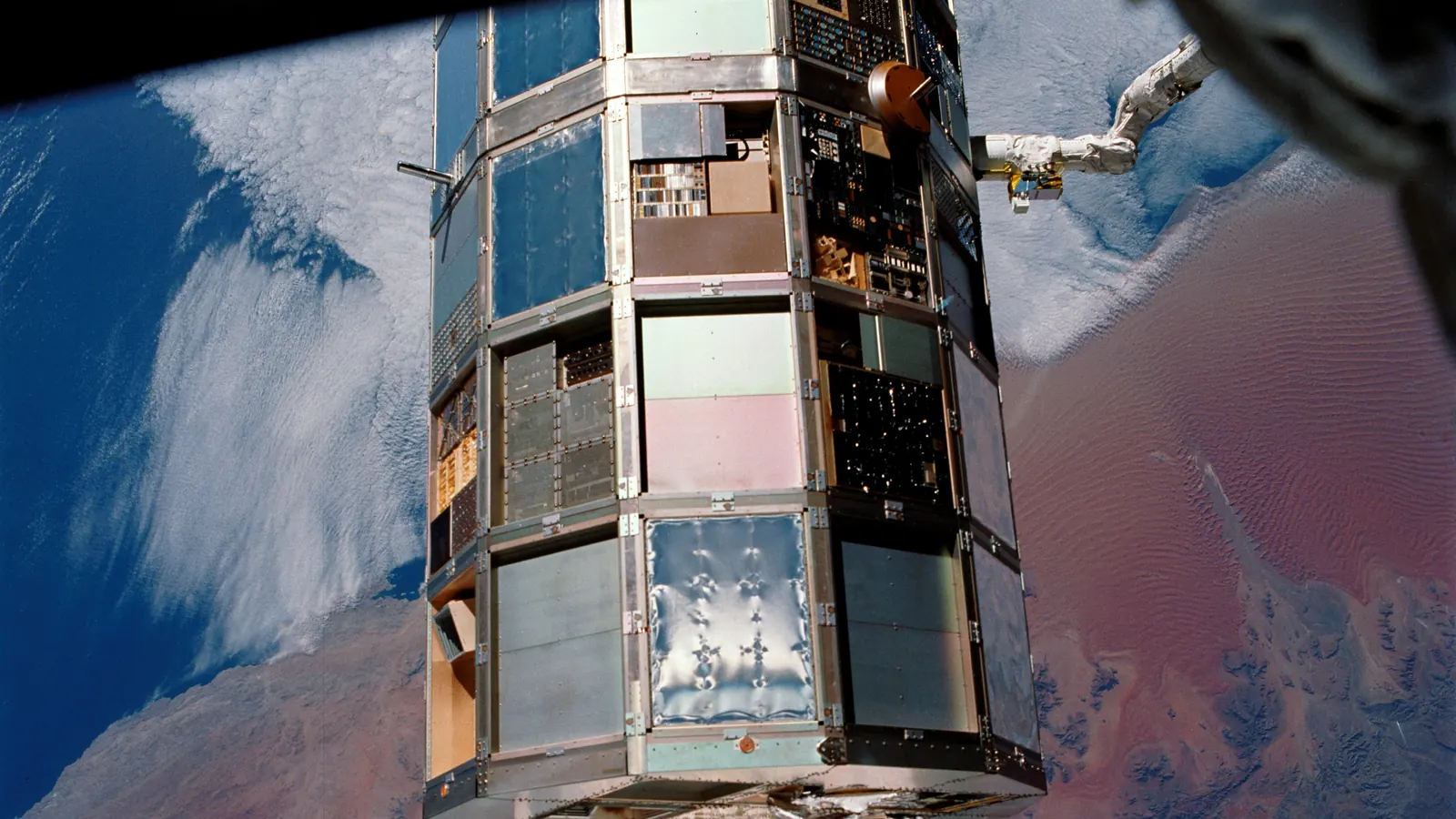 NASA's Long Duration Exposure Facility, a 12-sided satellite covered in experiment trays, held by space shuttle Columbia's robotic arm over the Namib Desert during its retrieval in January 1990