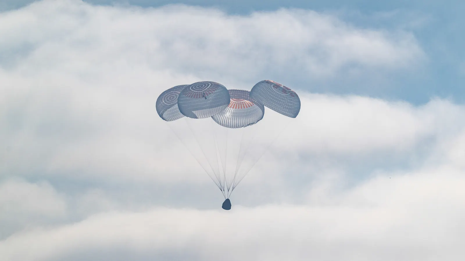 The Crew-12 Dragon capsule Freedom hanging beneath four open main parachutes against white clouds moments before splashing down in the Pacific Ocean off California on October 8, 2026