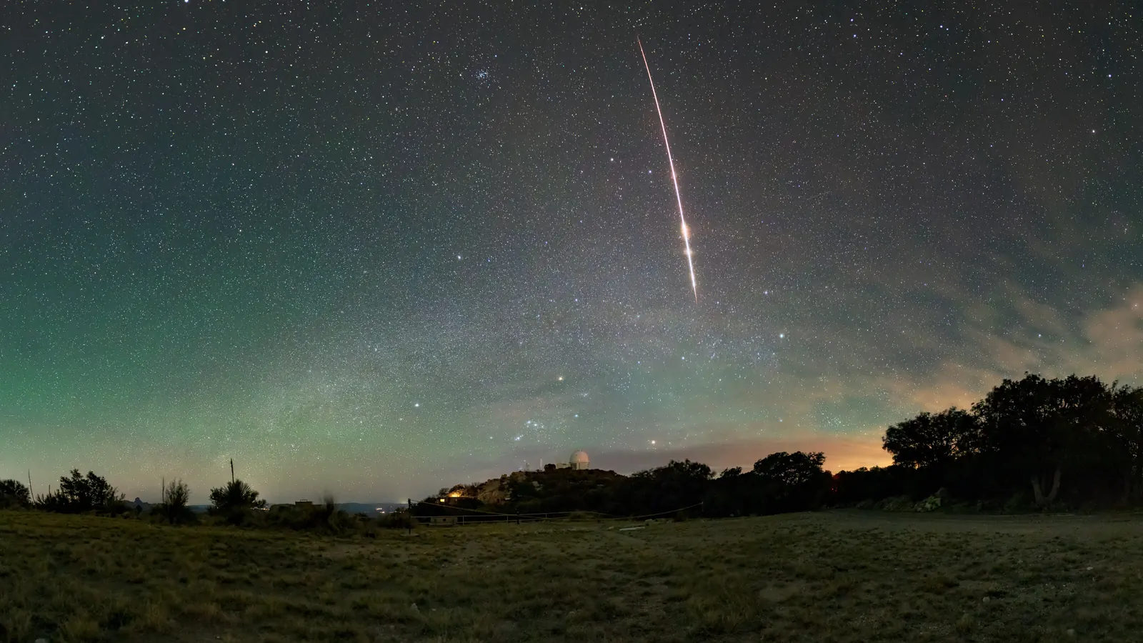 A bright fireball streaking down a sky full of stars above Kitt Peak National Observatory in Arizona, with the constellation Orion low over a white telescope dome and the Pleiades near the top of the frame