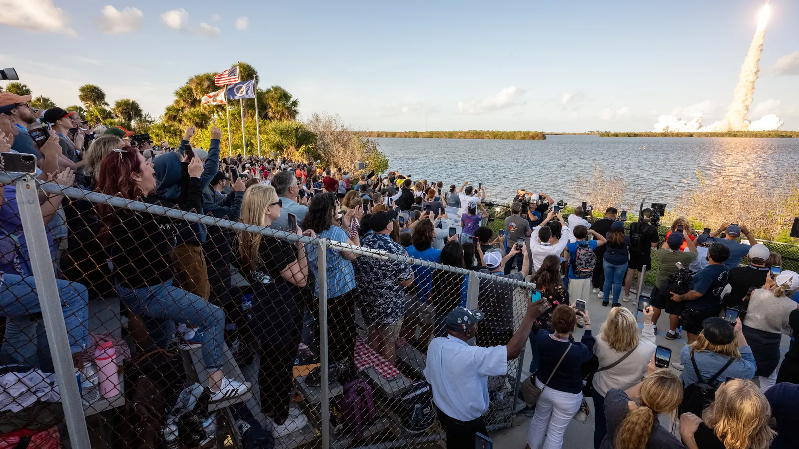 Crowds packed behind a fence by the water watch NASA's Artemis II rocket climb away on its smoke trail on April 1, 2026