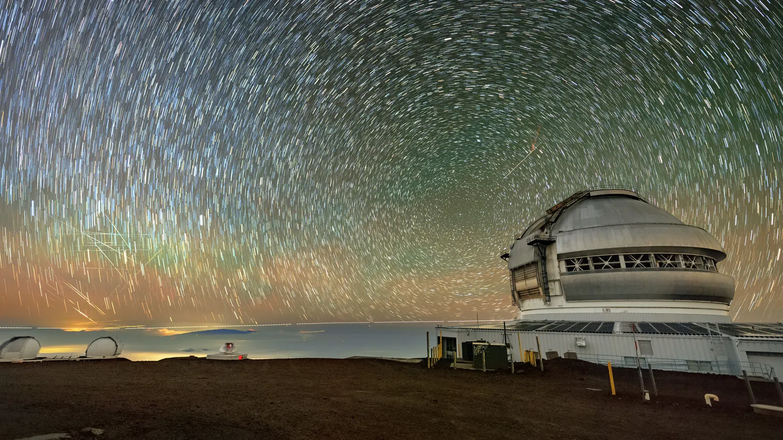 Star trails circling over the Gemini North telescope dome on Maunakea, crossed by straight satellite streaks low in the sky at left