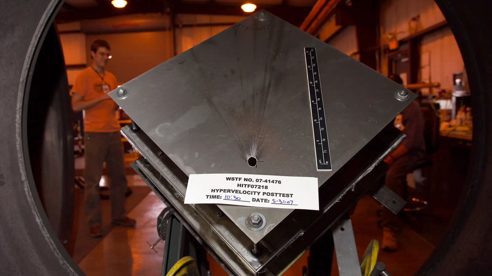 A metal test plate pierced by a small hole after a hypervelocity impact test at NASA's White Sands Test Facility, labelled "Hypervelocity posttest", with a ruler for scale and an engineer behind it