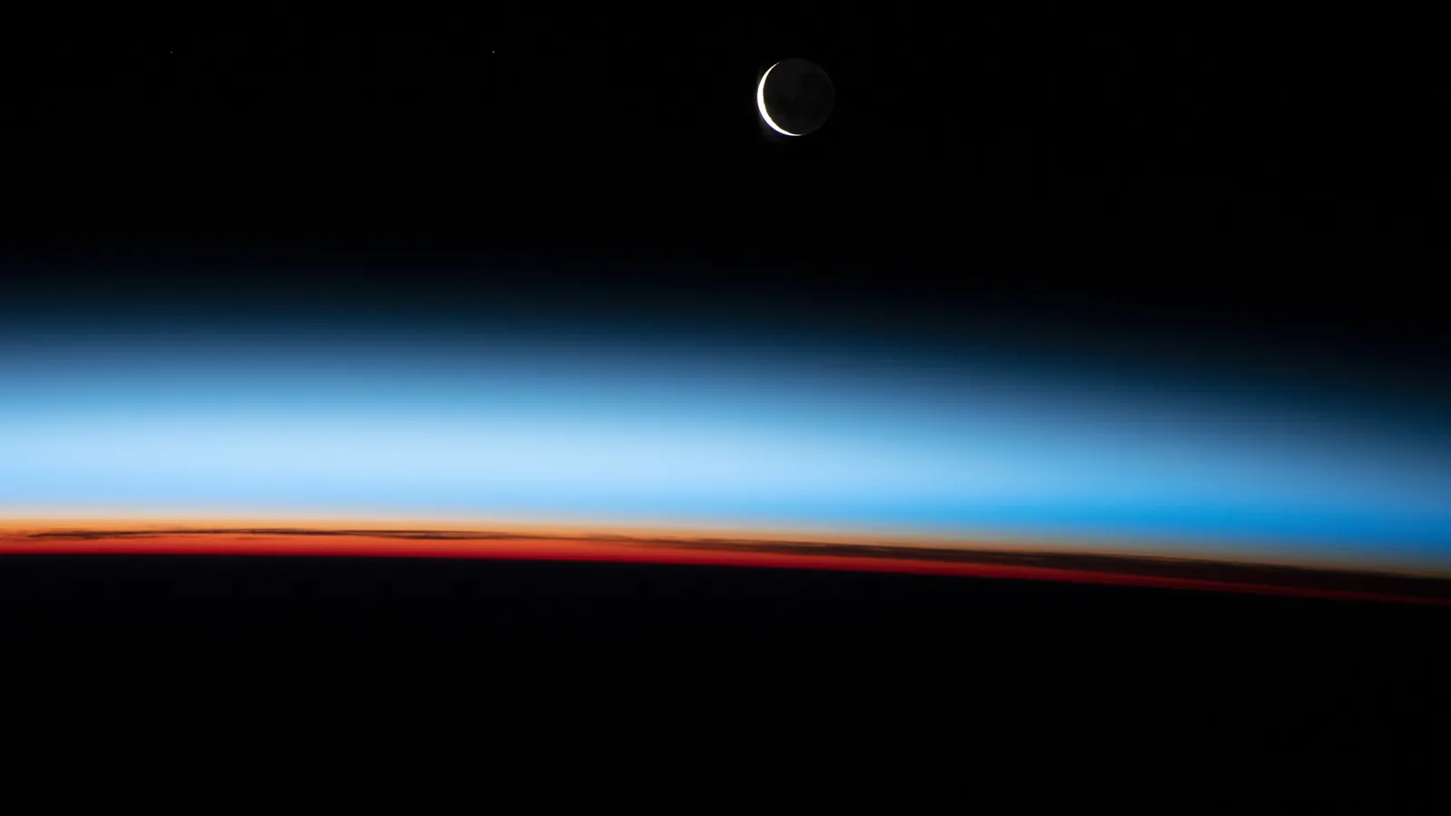 Earth's thin atmosphere seen edge-on from the International Space Station as glowing bands of orange and blue along the horizon, with a crescent Moon above